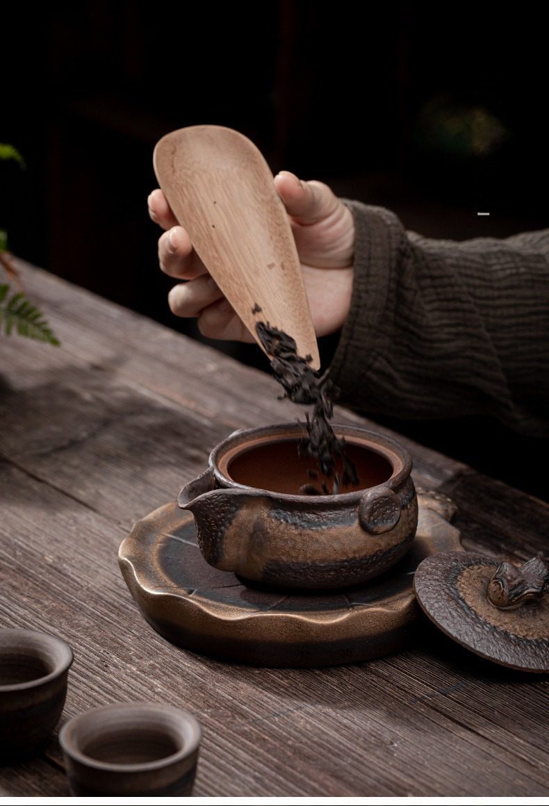古早燒木蓋鎏金手抓壺，南山居手作粗陶茶壺與木質茶則，展現古樸自然紋理，正將茶葉緩緩置入壺中，質感深沉細膩。