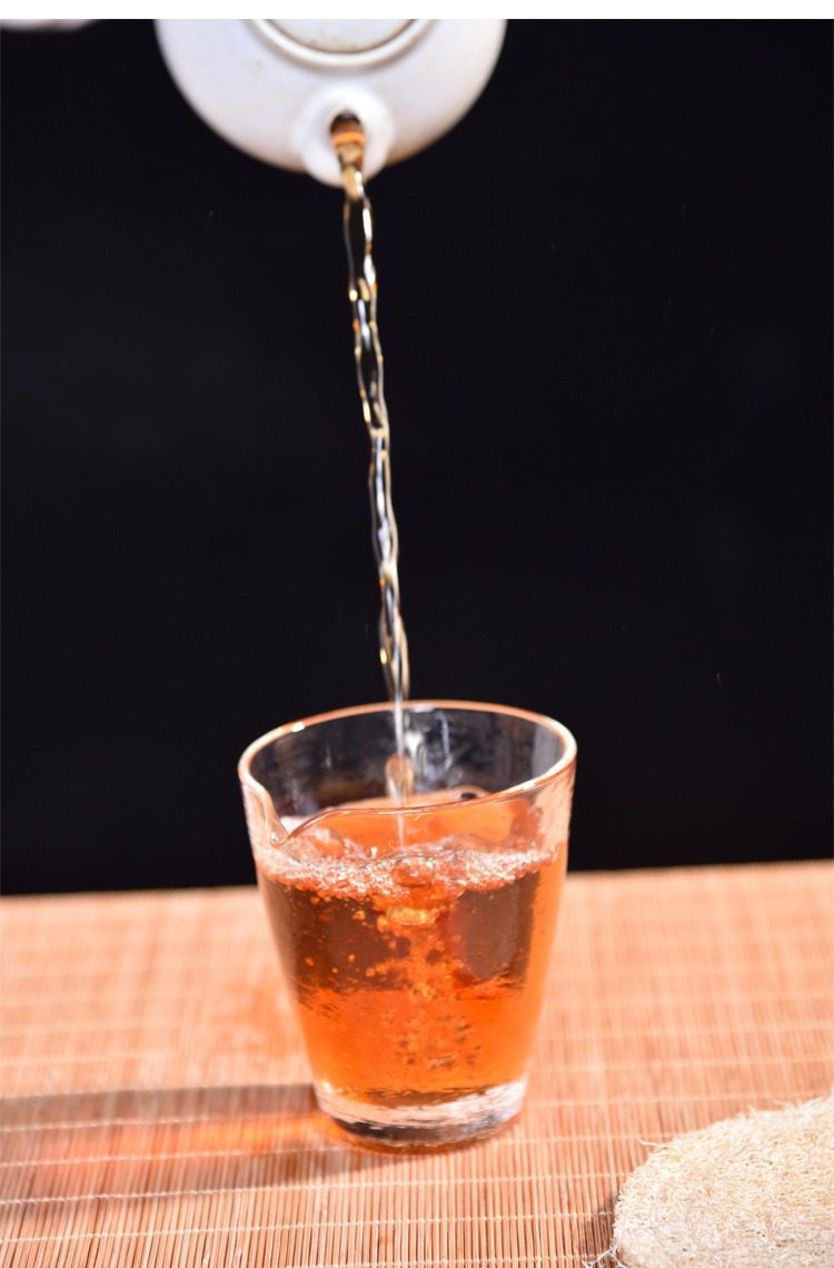 水波紋錘目玻璃公道杯，南山居透明玻璃公道杯，盛裝琥珀色茶湯，茶水自白色陶瓷茶壺優雅注入，背景為竹席與自然茶具擺設。