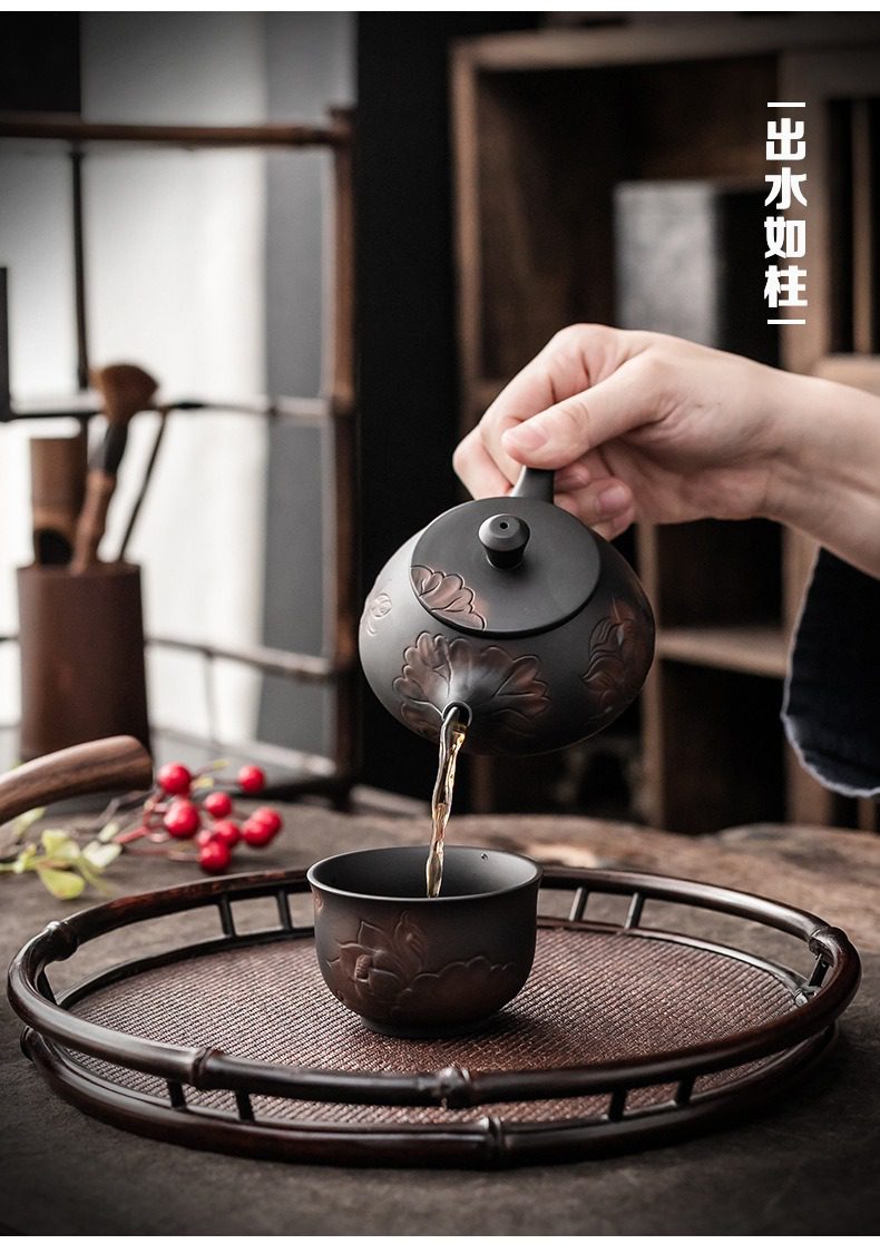 紫陶清溪蓮香茶壺，南山居手工雕刻蓮花紫砂壺，壺身呈圓潤古樸的黑色，配以浮雕蓮花圖案，搭配同款茶杯與竹編茶盤，正在優雅注茶。
