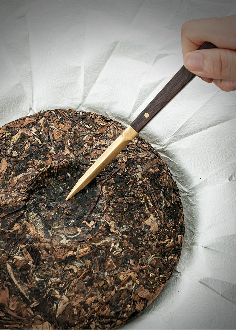 黑檀木茶道六君子杯墊套裝，南山居精緻木柄黃銅茶針，正輕巧撬開一片熟普洱茶餅，展現專業開茶技巧與茶具美學。