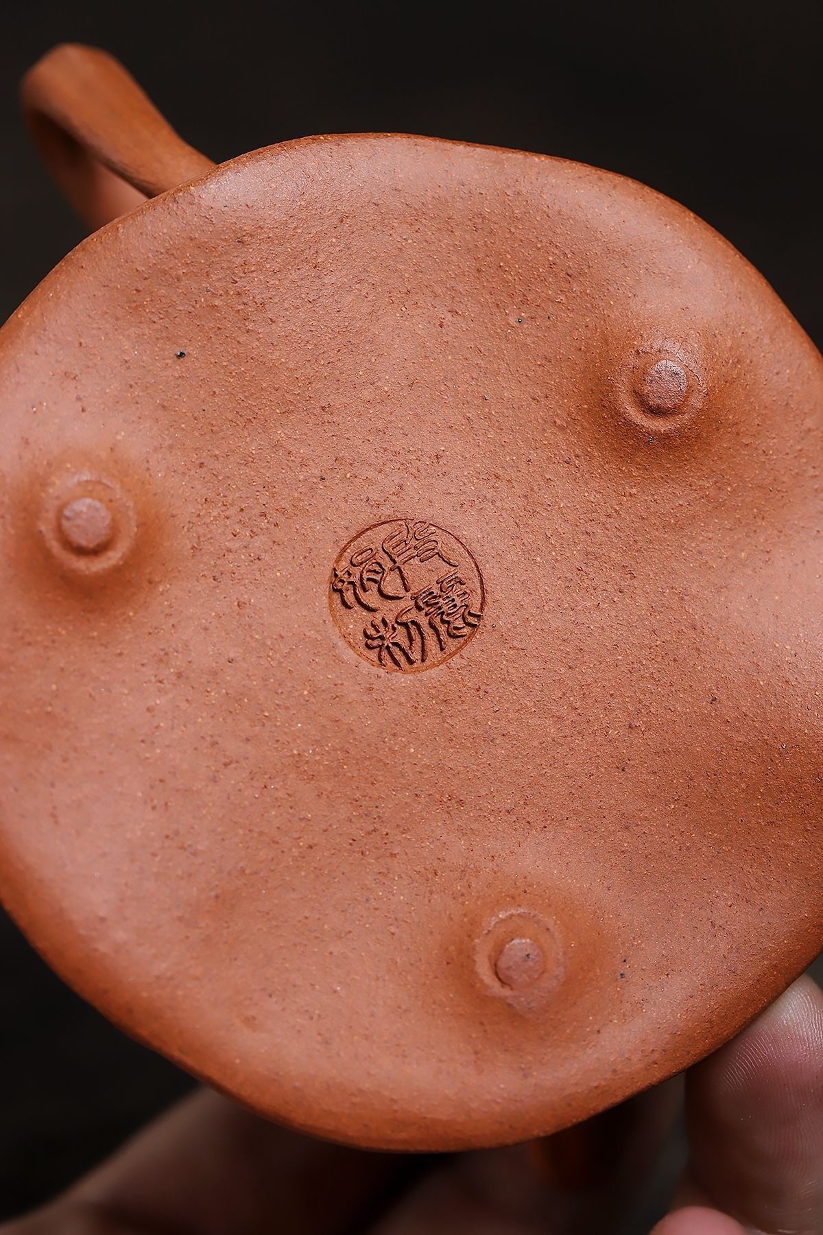 宜興紫砂壺原礦黃降坡蓮蓬仿生壺(140ml)，南山居紫砂壺底部特寫，展現溫潤朱泥泥料的細膩質感，中央刻有清晰藝師印款與三足設計。