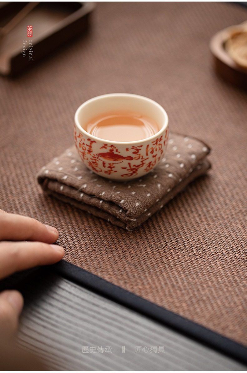 百圾碎蘇打釉品茗杯，南山居精緻手繪陶瓷茶杯，杯身飾有吉祥鯉魚與古典文字圖案，盛裝琥珀色茶湯，置於棕色斑點茶墊與編織茶席之上，呈現雅緻茶道氛圍。