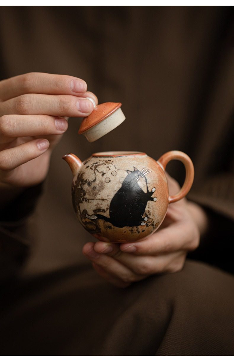 日式志野貓柴燒茶壺，南山居手工柴燒陶壺，壺身繪有生動黑貓圖案，泥色溫潤，造型圓潤雅緻，是獨特的品茗茶具。