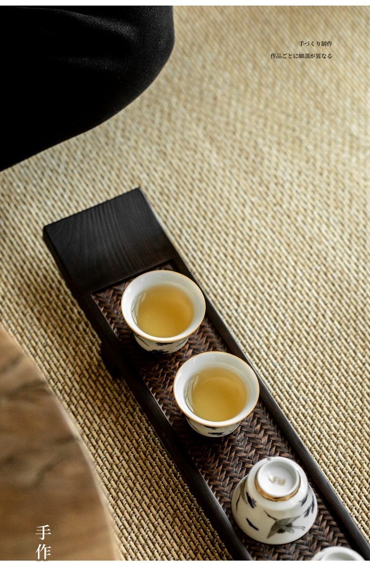 桐木高腳長杯架，南山居精緻陶瓷茶杯，杯身繪有淡雅水墨圖案，搭配木質藤編茶盤，盛裝金黃茶湯，展現品茗雅趣。