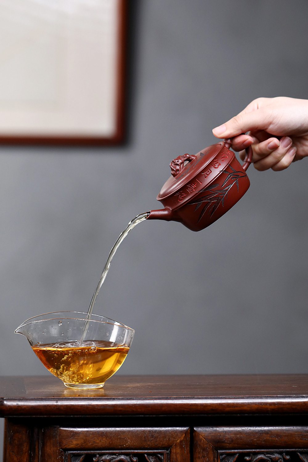 宜興紫砂壺原礦龍血砂太湖石竹韻壺(190ml)，南山居手工朱泥竹節壺，壺身刻繪清雅竹葉與詩文，壺蓋鈕為竹結造型，正將金黃茶湯緩緩注入公道杯，展現茶道美學。
