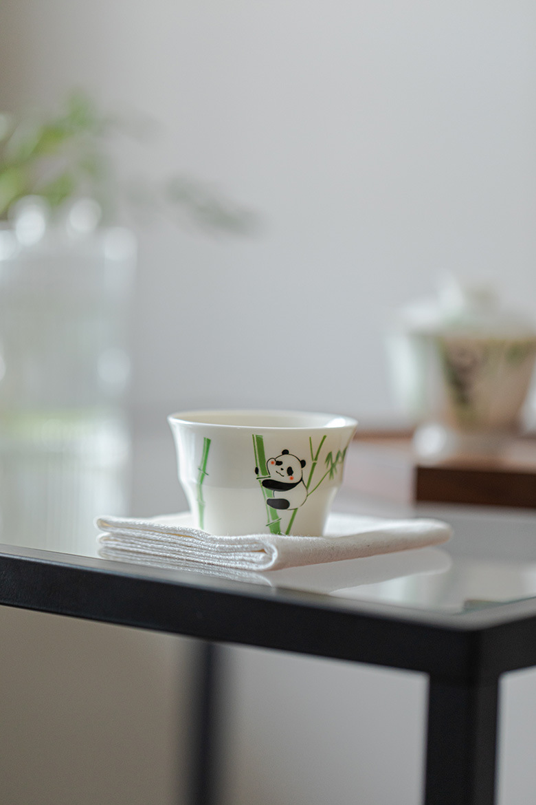 玉泥白瓷 － 竹節杯，南山居白瓷茶杯，繪有可愛熊貓攀爬竹林圖案，置於潔白茶巾之上，盡顯童趣禪意。